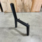A black metal chair frame or table base with a square backrest and horizontal support beam, photographed on a concrete floor in front of a wooden wall.