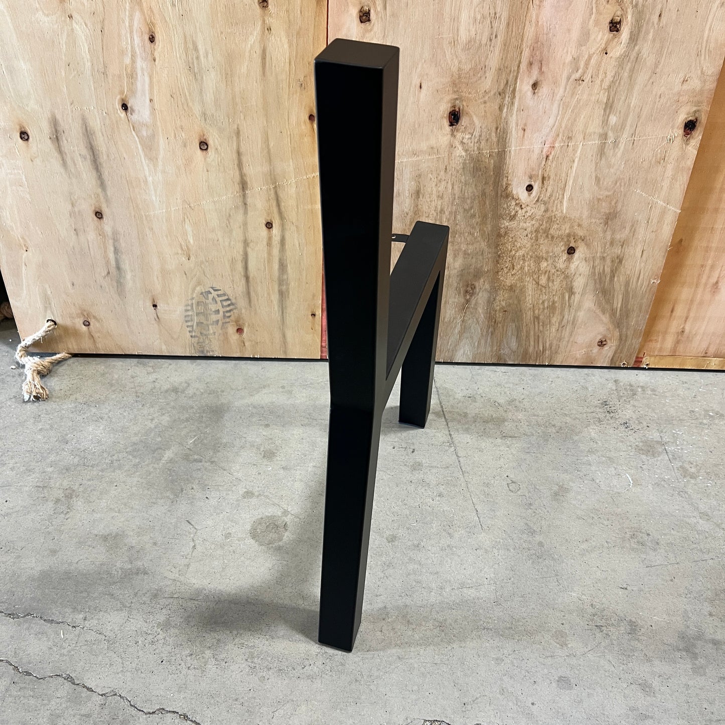 A modern black metal chair with a minimalist geometric design, standing on a concrete floor in front of a plywood wall.