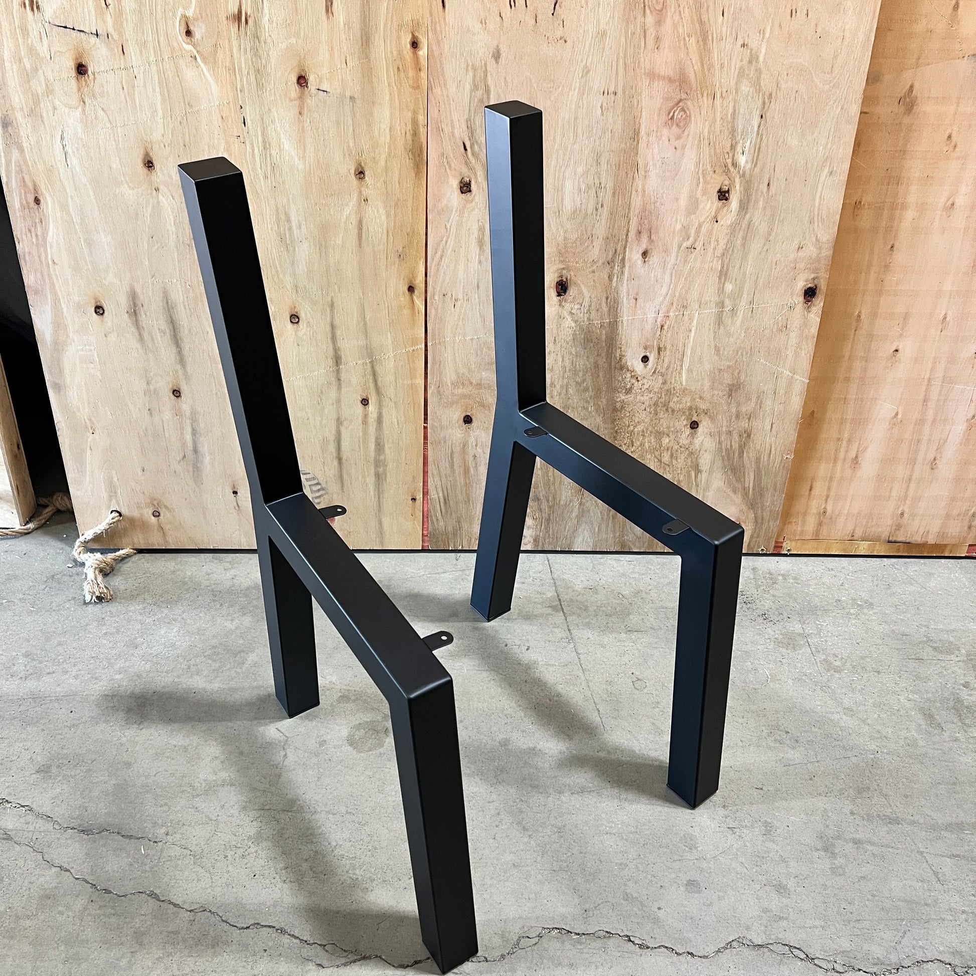 Two black metal table legs or base frames standing upright on a concrete floor in front of a plywood backdrop.